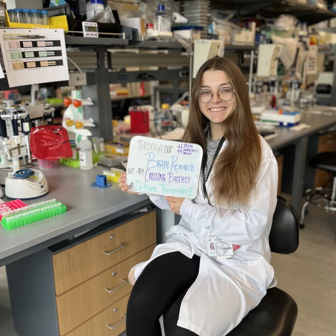 Woman sitting in a lab holding a sign that says, “Through our brain research we are crossing barriers to deliver therapeutics! #BrainAwarenessWeek”