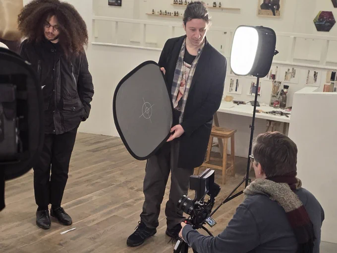 Three people on a practice film set in an art gallery space. One standing person holds some equipment while another crouches down with a camera. There is a stage light between them
