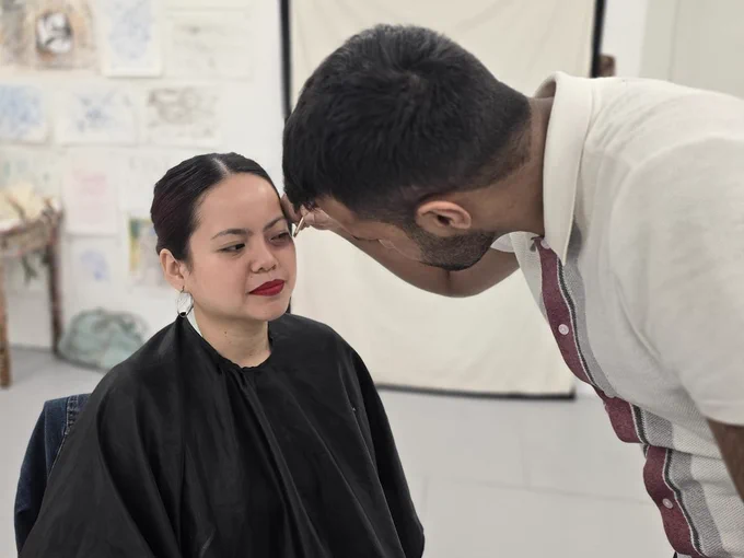 A person in a black smock sitting down having make-up applied by another person standing over them.