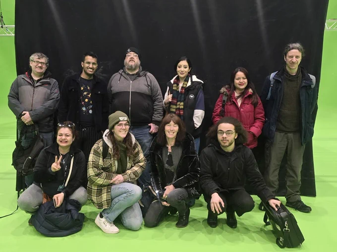 Ten people pose for a photo in a green screen studio in front of a black curtain. Four crouch in a line at the front while six stand behind.