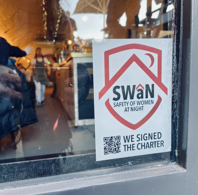 The SWaN sticker in the window of Mangos in Exeter, looking into the cafe bar with warm lights and someone working in an apron, white t shirt and jeans.