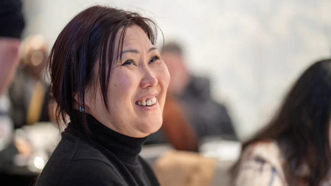 The image shows a woman in a black turtleneck jumper smiling, and looking off camera.