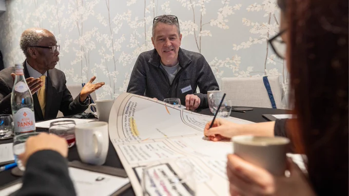 The image shows two men on one side of the table and a woman on the other. One man is talking, the other man is looking at the paper upon which the woman is writing.