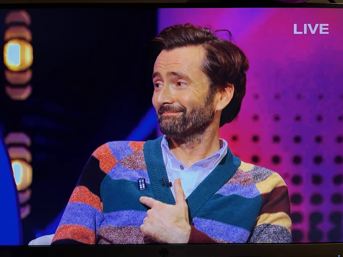 David Tennant pointing to a pin on his cardigan 