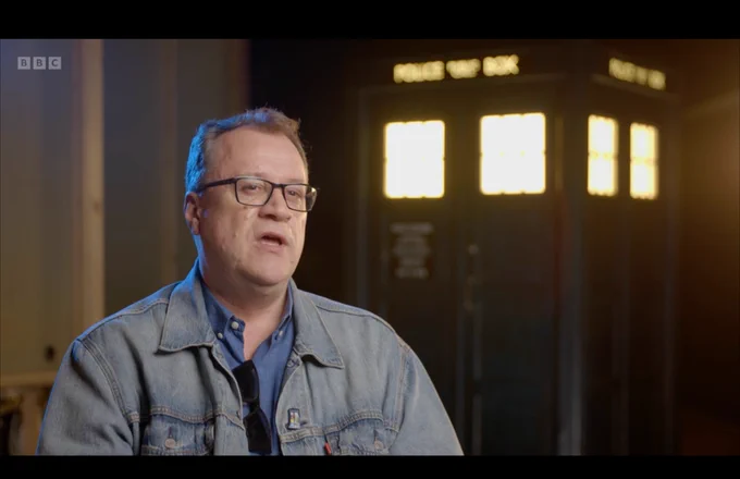Russell T Davis sitting in front of the Tardis, he is wearing a small rainbow pin on his lapel 