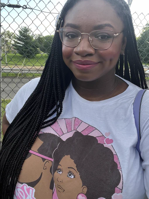 A photo of myself standing outside in front of a fence. I’m smiling and wearing a shirt displaying a Black version of Barbie and Ken.