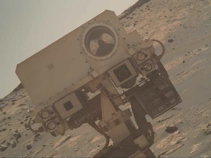 A beige-hued photo shows the "head" of the Curiosity Mars rover. It is one of many images the the rover took that created a mosaic in November 2021. The rover's rectangle-shaped head is off-center with the Martian horizon angled to the left along with it. The Martian surface takes up three-quarters of the background, showing a rocky and dusty terrain. The rover's rectangular head has a big circle to the right that is a camera. Below the rectangle are two square shapes and wires connecting to the neck of the rover.