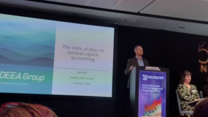 Man in suit speaks at podium, woman in green sits to his left, screen reads: IDEEA Group, the state of play on Natural Capital Accounting
