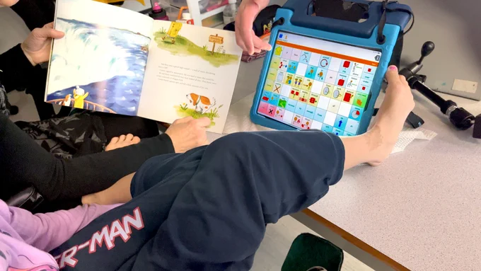 A student's leg propped up on a table, foot touching a device with a TouchChat communication screen. Beside someone holds up a picture book.