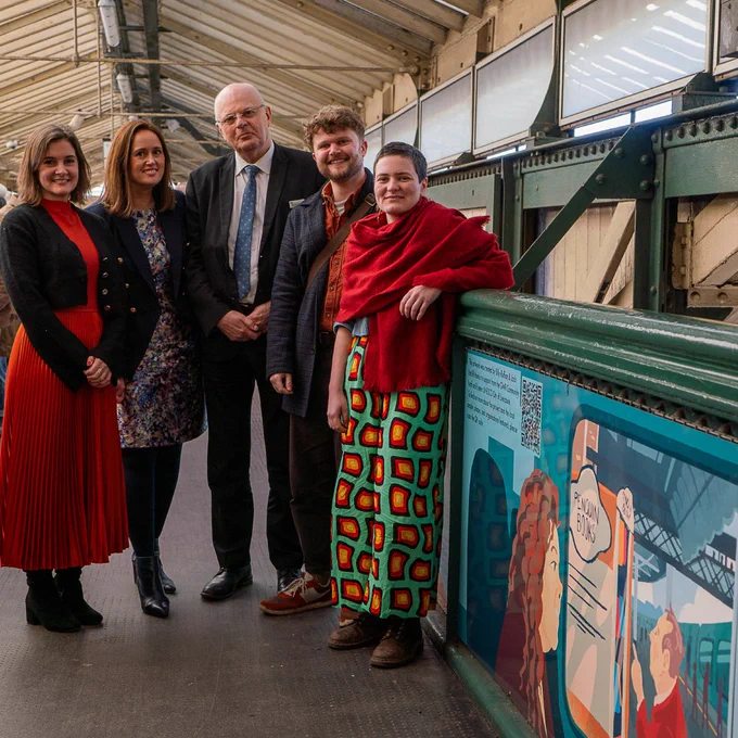 A group of five people stand on the footbridge of Exeter St David's Train Station next to a large panel of artwork depicting the station and the Penguin Books Vending Machine.