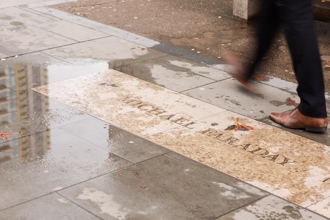 Photograph of a paved floor with 'MICHAEL FARADAY' engraved on it. In a puddle to the left is the reflection of a tower block and to the right a person's legs with black trousers and brown shoes can be seen walking past, one blurred as it moves.