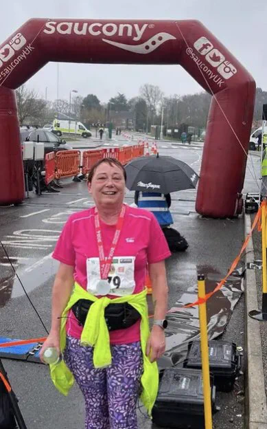 Image: Tracey smiling with the finish line behind her, Tracey is wearing a pink T-shirt, a bright yellow top tied around her waist, running trousers and has a fab medal around her neck.