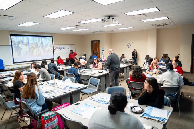  View of the entire room of Project TLC participants at tables and the projector screen picturing images of students