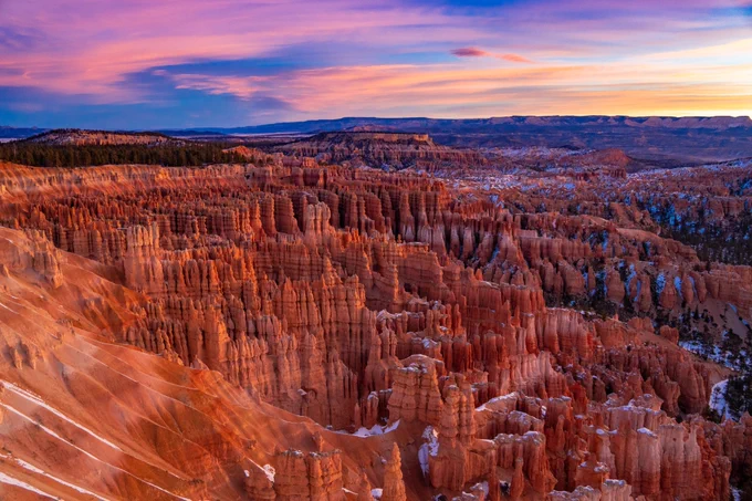 As the sun rises, pink, orange and yellow colors fill the sky over a landscape with tons of geologic formations called hoodoos. 
