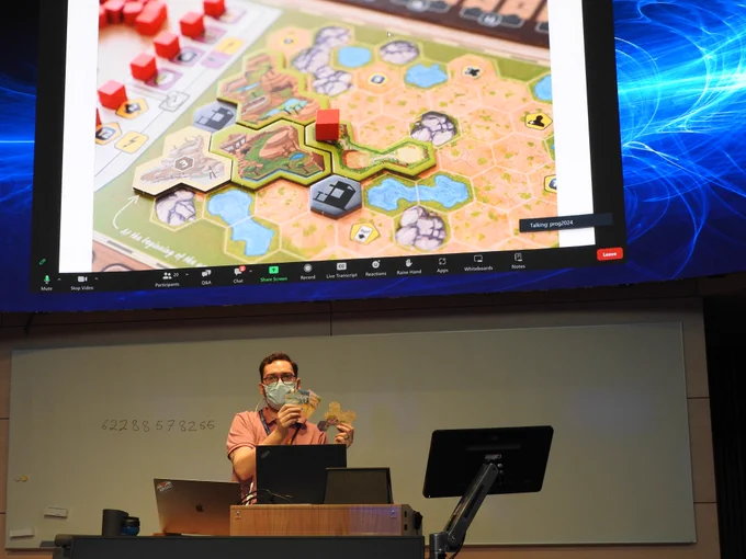 Photo of a presentation in the PAI track. The speaker is holding parts of board game that was used in the evaluation of their approach.
