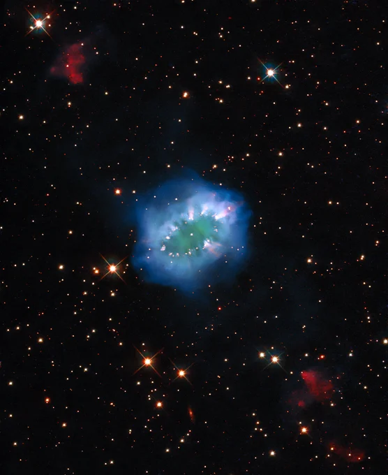 A small, bright green region of gas is surrounded by a ring of glowing cosmic material, concentrated in pale clumps that resemble diamonds along a necklace. The rest of the image shows black space dotted with several bright stars and a couple small regions of dark red gas.