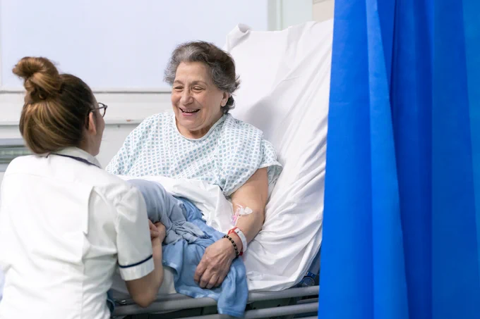 Photograph of an elderly female patient sitting up in a hospital bed and talking to a member of staff who is knelt at her bedside.