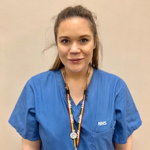 Portrait of Georgina, a nurse at Imperial College Healthcare. Georgina is wearing her blue, NHS hospital scrubs and an NHS lanyard. She is smiling towards the camera.