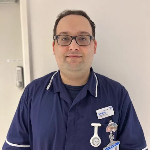 Portrait of Jonathan, a nurse in charge at Imperial College Healthcare NHS Trust. Jonathan is wearing a navy blue nurses uniform and smiling towards the camera. He is wearing black glasses.