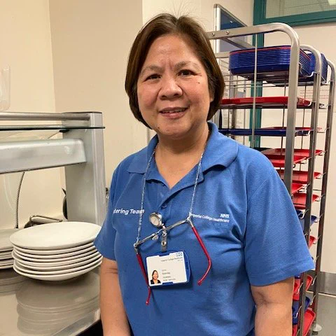 Portrait of Edna, a ward host at Imperial College Healthcare. She is standing in a hospital kitchen with a food and drink trolley behind her.