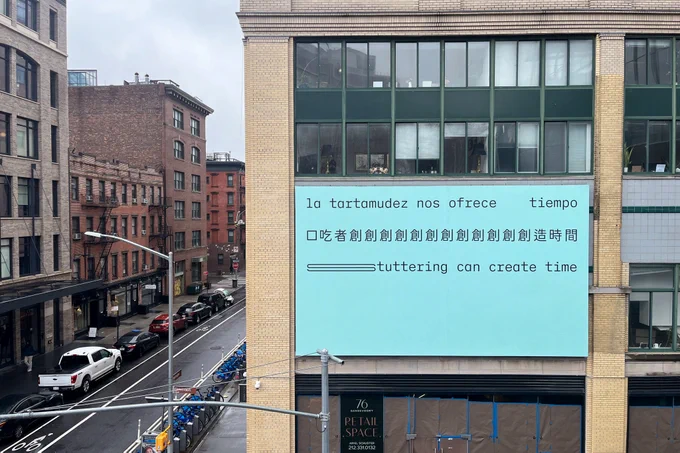 A photo of a billboard on a New York City building. Three lines of black text appear on a light seafoam green background: ‘La tartamudez nos ofrece tiempo, 口吃者創創創創創創創創創創創造時間, Stuttering can create time.’ The text is in a sans serif typeface organized in three straight lines within the top half of the composition. The bottom half of the composition is empty emphasising a sense of pauses, silences and expectations. The text is all lowercase and lacks punctuation, giving a casual and informal almost text message-like feel. Each line of text represents a form of stuttered speech: blocks, repetitions and prolongations. The first line in Spanish translates to “Stuttering offers us time.” The space between “us” and “time” represents a block. The second line in Chinese translates to “People who stutter create time.” The first characters in the Chinese word for “create” are repeated. In the third line in English the “s” in “stuttering” is stretched horizontally to represent prolonged sound.