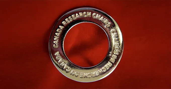 A silver circle broach with the words "Canada Research Chair" inscribed. The broach is pictured on a red background. 