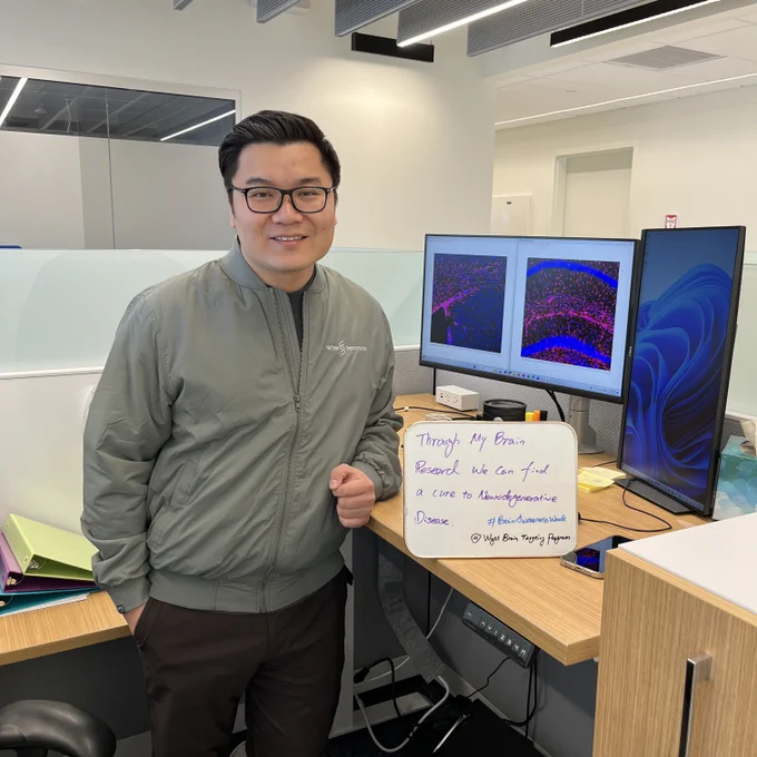 Man standing next to a desk with a computer showing microscopy images. He is next to a sign that says, “Through my brain research, we can find a cure to neurodegenerative disease. #BrainAwarenessWeek Wyss Brain Targeting Program.”