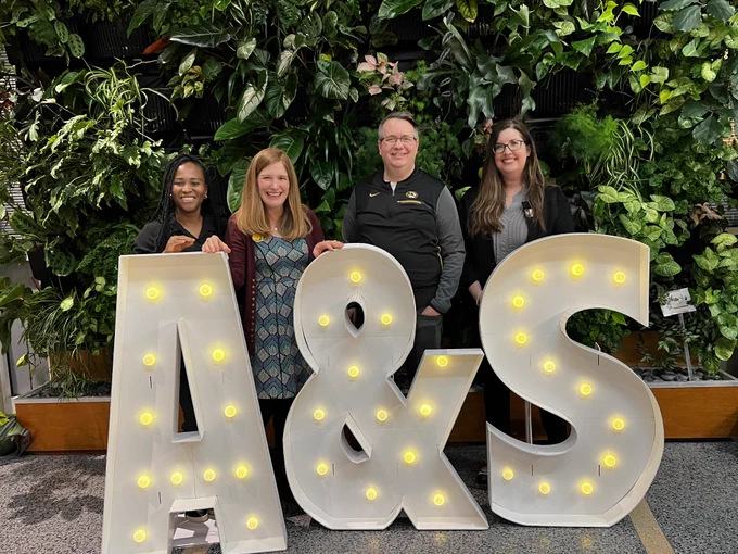 Dr. Horstman, Dr. Warner, Dr. Behm-Morawitz, and PhD Candidate Blessing Okafor smile for the camera behind large white "A&S" marquee letters.