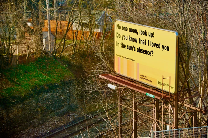 A billboard on a sunny winter day, with leafless trees and some vegetation. Train tracks intersect the photograph. The billboard, mostly in yellow, features a haiku, credited to Jason Livingston and Phoebe A. Cohen: “No one noon, look up! Do you know that I loved you in the sun’s absence?” Vertical lines in gradients of orange and yellow are next to the hashtag #InthePathofTotality, and Squeaky Wheel’s logo.