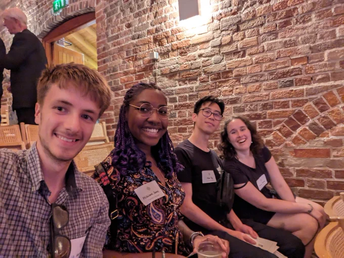 From left to right: Will Harmer, Millicent James, Alex Tay and Emily Hazrati sat in a row on wooden seats next to a brick wall; ready to see National Youth Choir 18-25, London Youth Choirs and The Swingles in Snape Maltings Concert Hall, Suffolk (photo taken August 2023).