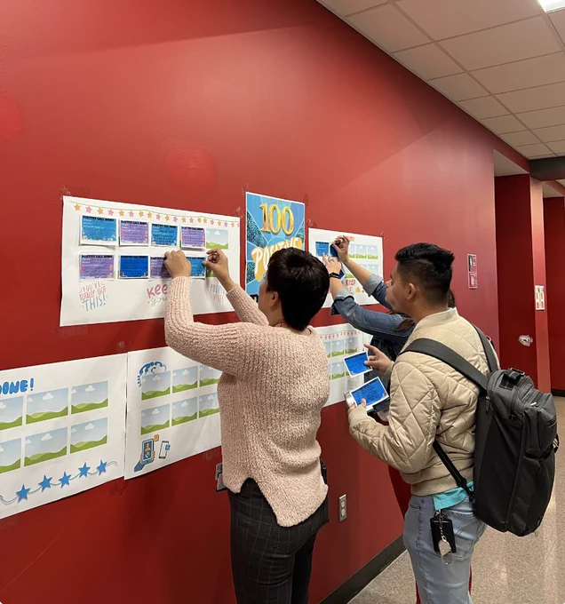 In a hallway hangs a poster that says "100 positive calls." Three adults in the hallway add small pieces of paper to a larger chart tracking phone calls.