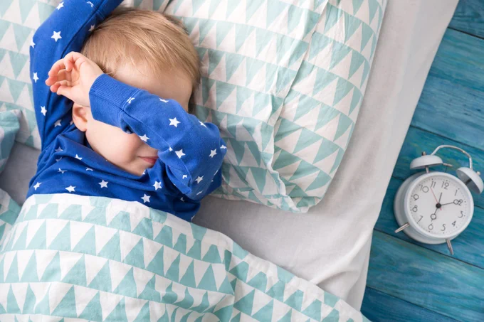 A sleeping child in bed next to an alarm clock.