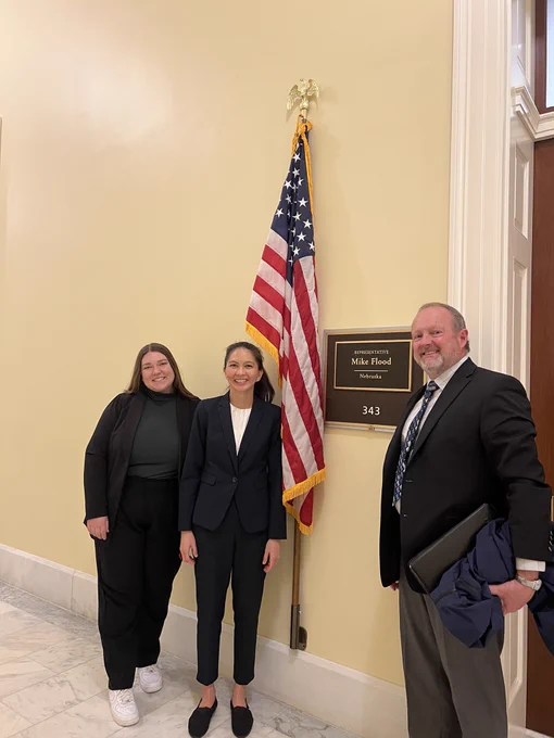 Three people standing outside representative Mike Flood’s office, smiling and posing for a picture. They are standing near a flag and a plaque with the Rep. Flood’s name and state (Nebraska).
