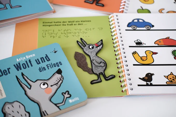Originalbuch "Der Wolf und die Fliege", daneben aufgeschlagenes taktiles Kinderbuch.