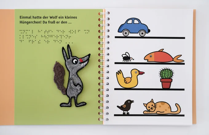 Aufgeschlagenes Buch: Links Text in Brailleschrift und Großdruck mit Wolf, rechts das Regal mit Katze, Vogel, Kaktus, Ente, Fisch, Fliege, Apfel und Auto. 