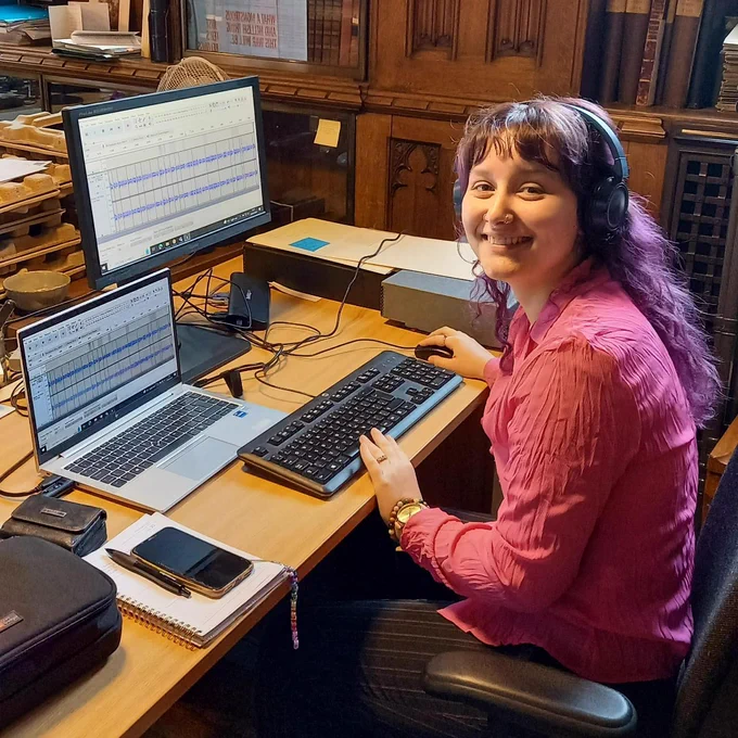 Matilda Seymour-Lewis editing audio at a desk using a computer. She is in one of the historic rooms of the Rylands Library.