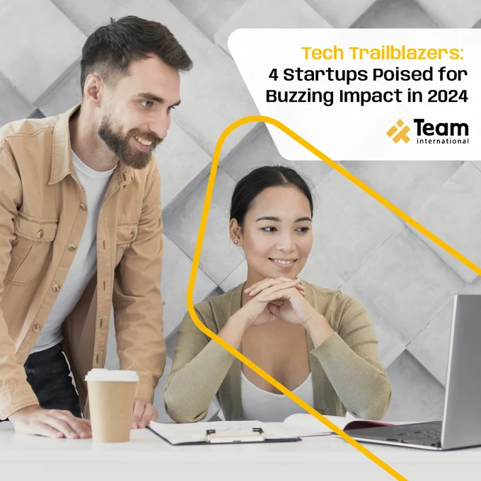 A man and a woman are watching a laptop, they are on a desk, and on it, there's a coffee cup and a notebook. In the right corner is the Team International logo