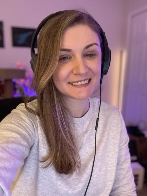 A selfie of Crev wearing a grey crew neck sweatshirt she has long brown hair and green eyes and is wearing a pair of black and grey bose headphones.
