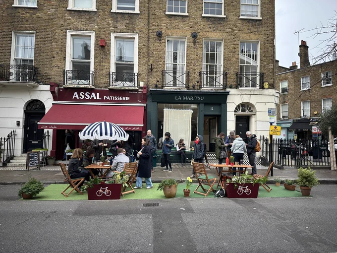 Parklet on green surface outside two shops. Wooden tables and chairs. Planters. People sat and stood chatting.