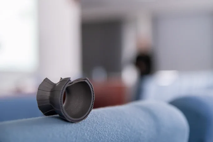 An additively manufactured part sits on a chair with a blurred background of the presenter in a conference room.
