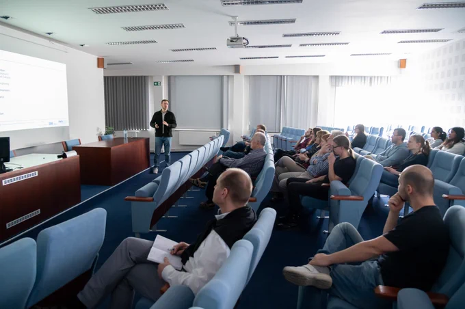 A presenter stands at the front of an auditorium giving a presentation to a focused audience.