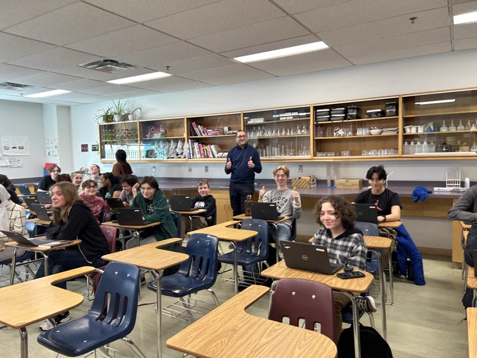 Des élèves utilisent des ordinateurs portables dans une classe, et leur enseignant se tient près d’étagères contenant du matériel de laboratoire.