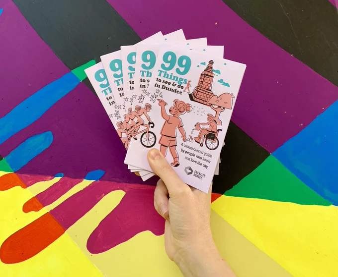 A hand holds up a fan of five copies of the 99 Things guide in front of a brightly coloured mural. The top of the mural is purple and black stripes moving into green and blue and yellow at the bottom.
