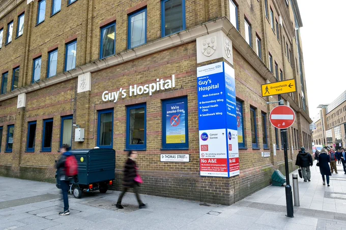 An exterior corner of Guy's Hospital, with a white sign bearing the hospital's name, and people walking by