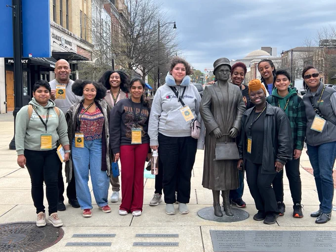 A group picture of the students in the DEI Common Read with two NOVA faculty members, Dr. Sabrina Ricks, Associate Professor in the Business and Public Services department at NOVA, and Ken Abrams, Assistant Professor in the Administration of Justice department, who served as chaperones. All are standing around a statue.