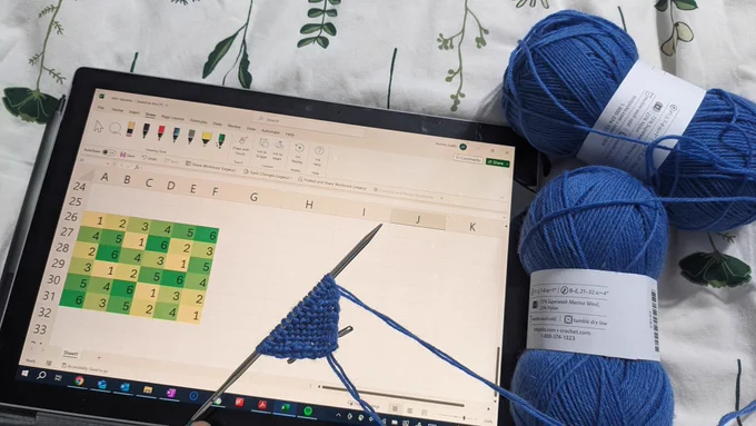 A photo of a tablet showing an Excel spreadsheet featuring a 6×6 Latin square. On top of the tablet is a knitted right-angle triangle, and to the right of the tablet are two balls of yarn.