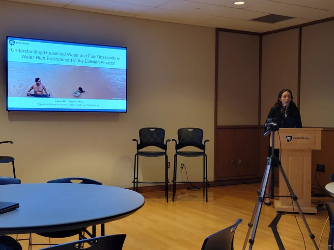 Dr. Lauren Broyals presenting her presentation titled Understanding household water and food insecurity in a Water-Rich environment in the Bolivian Amazon. The slide shows a man and a woman in a brown river and Lauren behind the podium. 