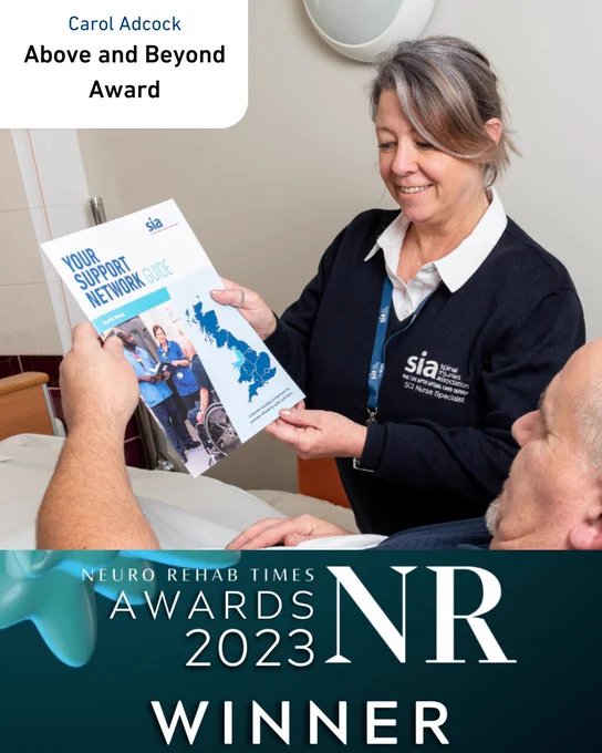 A picture of a woman smiling wearing an SIA jumper. She is showing an SIA brochure to a man in a hospital.

In the top left, text says: 'Carol Adcock. Above and Beyond Award'

At the bottom is a banner for the NR Times Awards Winners.
