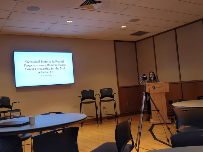 Speaker Brandi Gaertner behind the podium beside a slide reading, Geospatial patterns in runoff projection using random based forest forecasting for mid Atlantic US. 