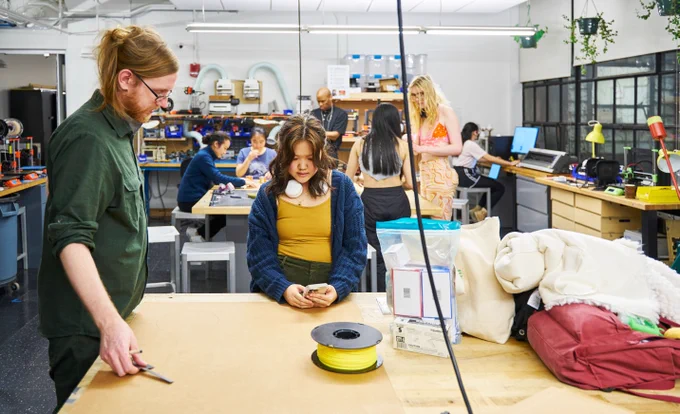 The photo depicts the N52 fabrication shop, a brightly-lit space with numerous miscellaneous tools on every surface. In the foreground, Bill McKenna, N52 instructor, assists a student. McKenna is wearing a green, collared shirt with sleeves rolled up to his elbows. He holds a small right-angle ruler. The student is a young woman wearing a yellow tank top and knitted blue cardigan. She is holding her phone as if to share an example photo or idea with the instructor. In the background are numerous other students seated at a table working on various tasks.